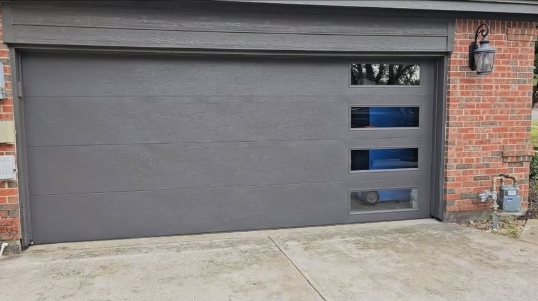 Gray flush-panel garage door with a vertical row of windows on a red brick home